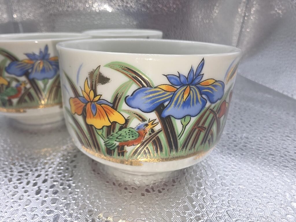 Close view of a porcelain yunomi showing blue and yellow iris flowers, a small green-and-orange bird and gold accent band, with a second cup blurred behind