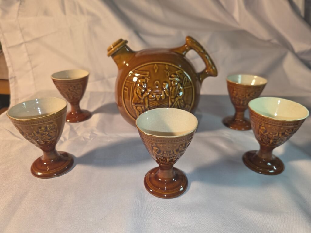 Brown glazed circular ceramic decanter with handle and cork at back, five matching footed goblets arranged in front; raised tavern-relief and crackle interior visible.