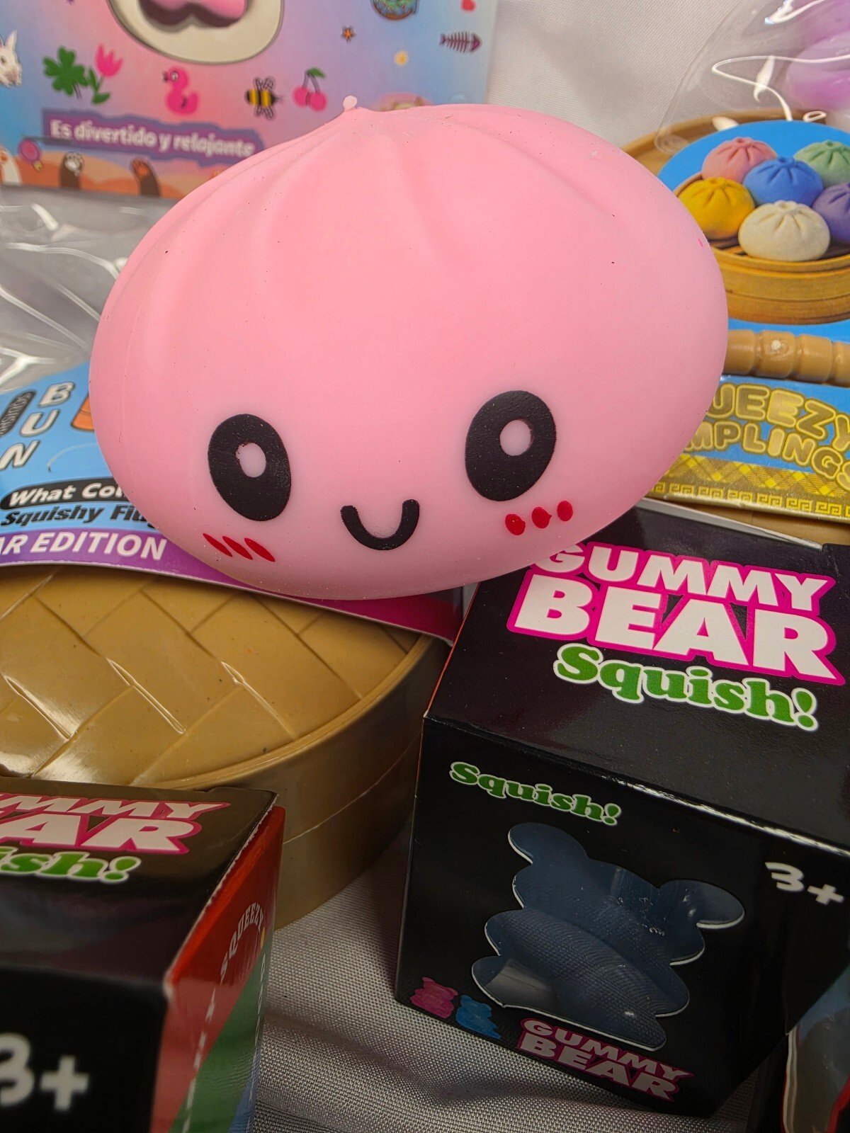 Front view of a pink smiling bun squishy with printed eyes and cheeks resting atop product boxes.