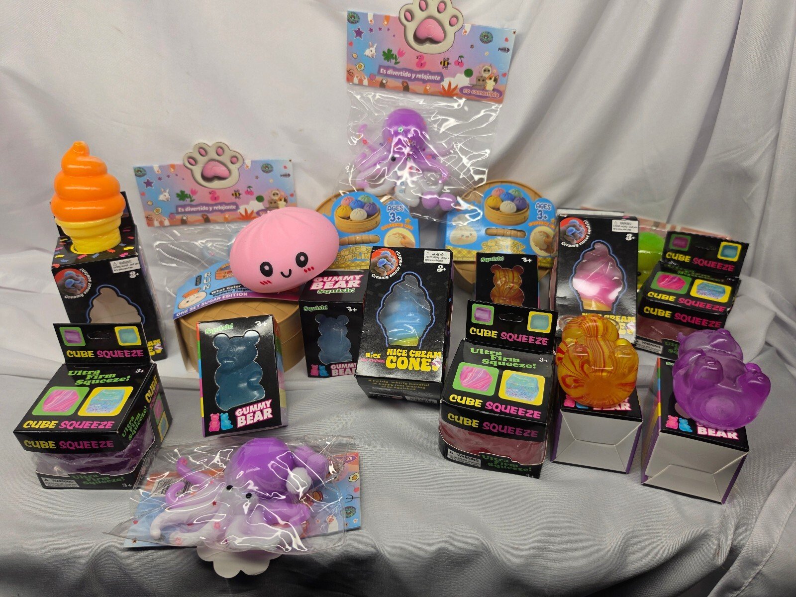Wide-angle group layout of assorted squishy toys and packaging arranged on gray fabric, featuring gummy bear and ice cream cone boxes.