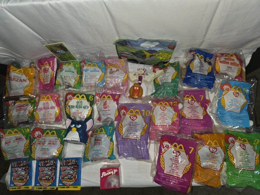 Top-down view of numerous sealed and loose McDonald's Happy Meal toy bags on white cloth, showing Desert Storm card packs, a Slinky Jr. box and a loose Furby figure