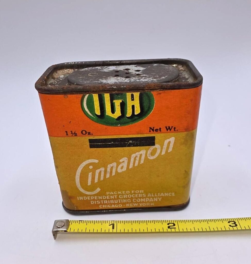 Three-quarter front/top view of IGA metal cinnamon tin showing orange-yellow label, '1½ Oz' marking, rusted shaker top, and measuring tape at base