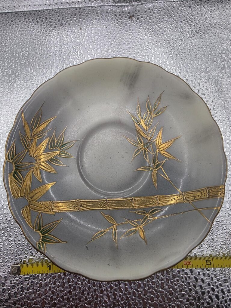 Close top view of gray porcelain saucer with gold bamboo moriage decoration and scalloped rim; measuring tape at bottom.