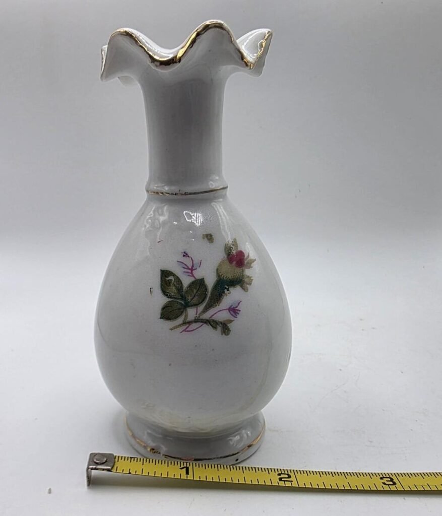 Front view of white porcelain bud vase with ruffled gold-trimmed rim and pink rose decal; measuring tape visible along base.