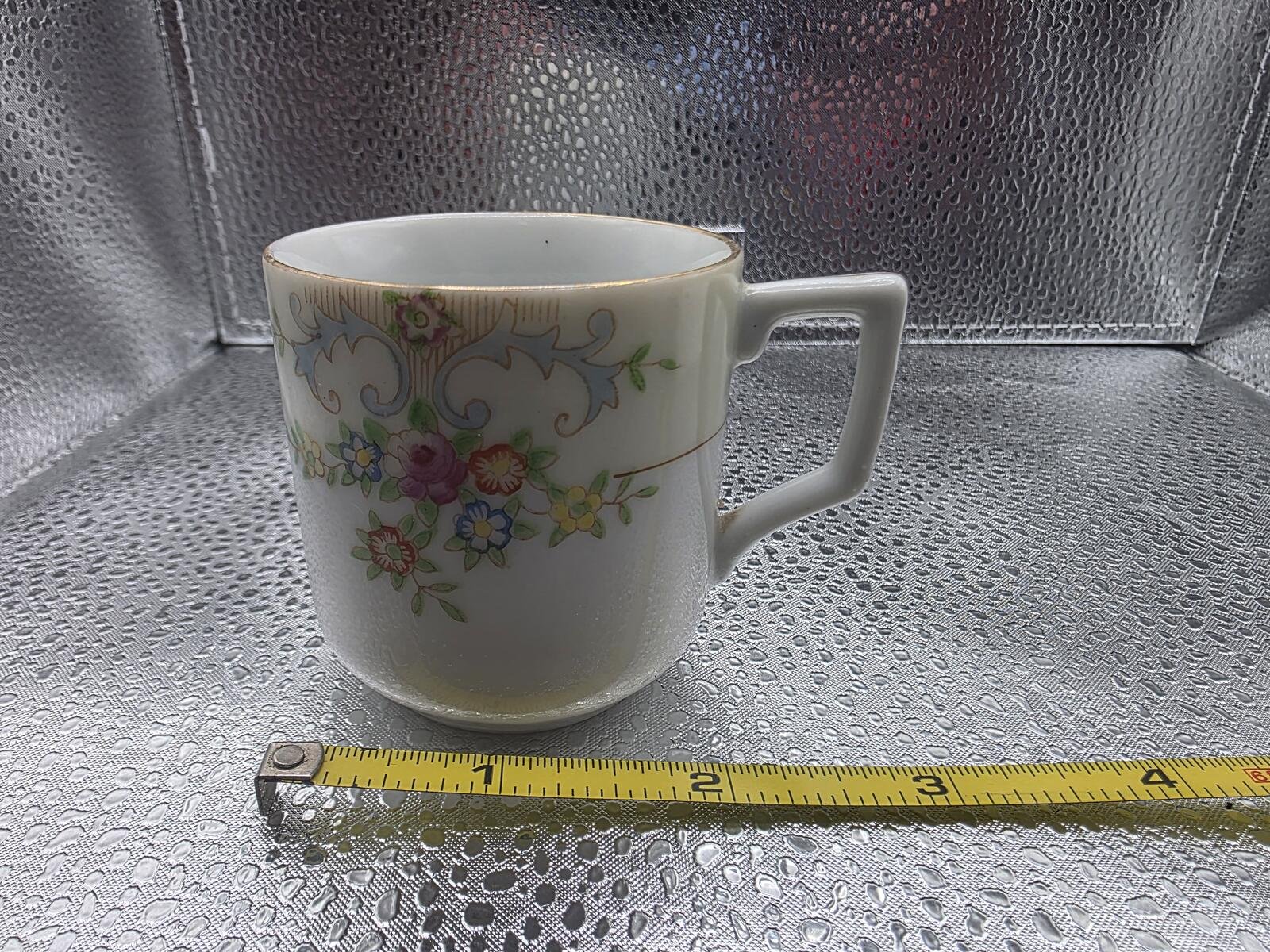 Porcelain demitasse cup, right-side handle view, hand-painted pink and blue floral band with gold rim; measuring tape at base shows about 3 inches