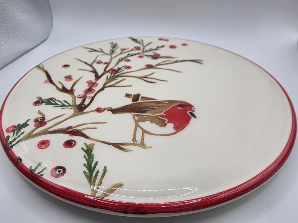 Close view of round glazed plate showing a hand-painted red-breasted robin on a branch with hawthorn berries and a red rim.