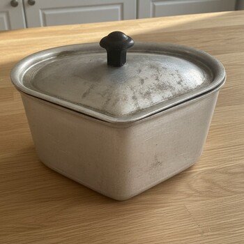 Top-front view of triangular aluminum covered pot showing lid profile, original rounded black knob, and scuffed, aged surface.