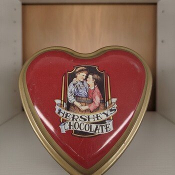 Front view of red heart-shaped Hershey's metal tin on shelf showing lid artwork, gold-toned rim and repeating gold 'H' logos on the sides