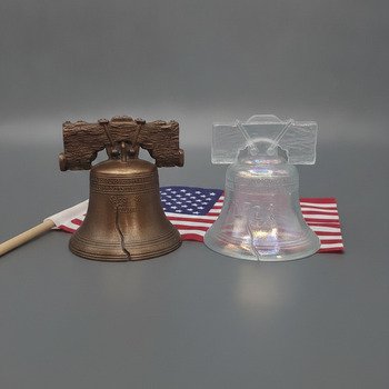 Square crop of two Liberty Bell replicas on gray surface: bronze-tone bell with textured beam beside translucent iridescent bell and small American flag partially visible.