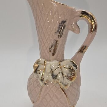 Close-up side view of pink ceramic pitcher showing handle, white leaf detail, gold trim and visible hairline crack.