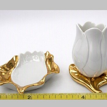 Close side view of white porcelain tulip bud vase on gold-tone leaf base beside matching gold-trimmed leaf-shaped dish.