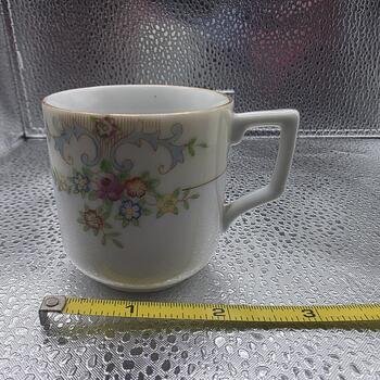 Front-side view of porcelain demitasse cup showing floral garland, square handle and gold trim with measuring tape at the base